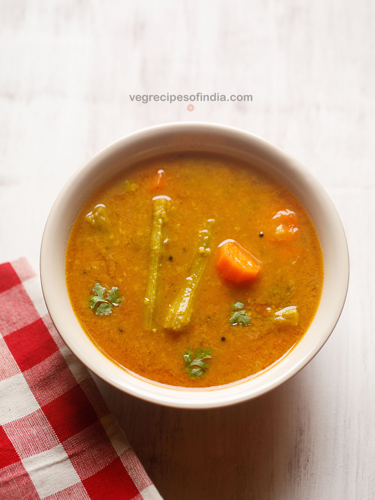sambar served in a cream bowl on a white board with a red & white checkered napkin on the side