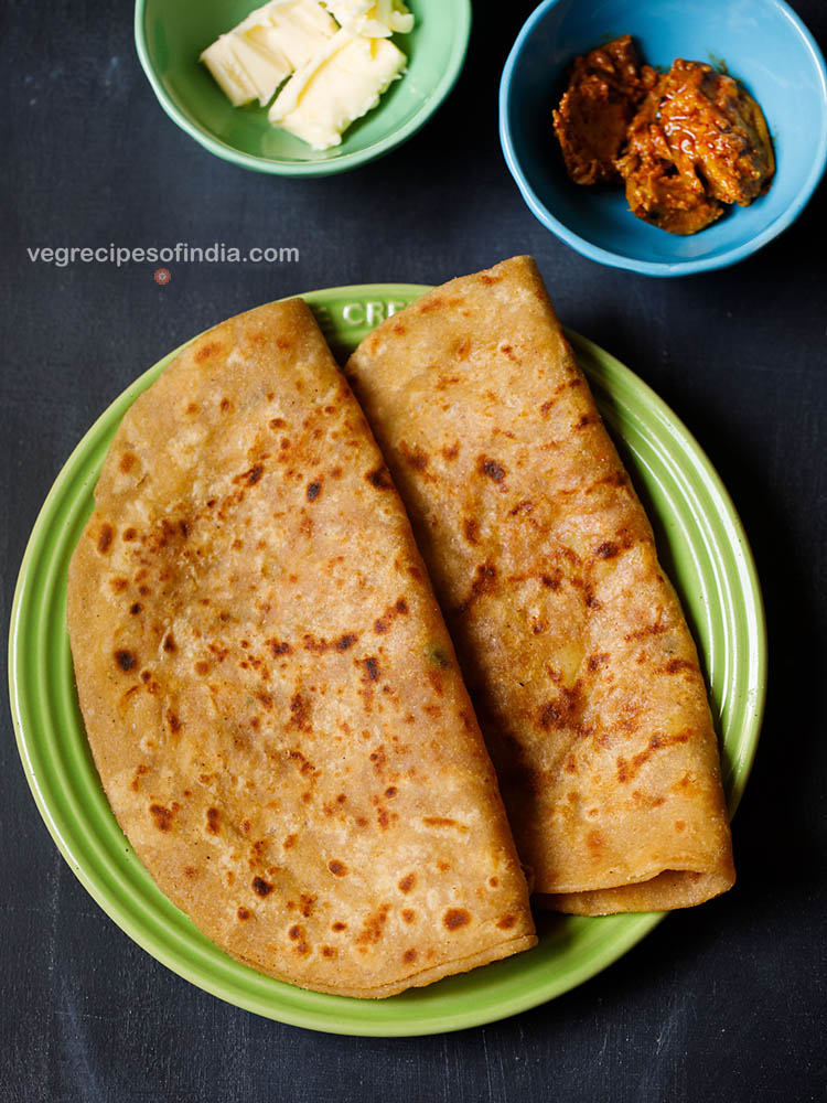 aloo ke paratha folded and placed on a green ceramic plate on a dark bluish board. sides of butter cubes and mango pickle served in two small ceramic bowls.