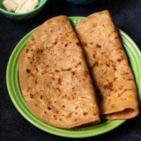 aloo paratha folded and placed on a green ceramic plate on a dark bluish board. sides of butter cubes