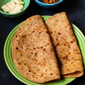 aloo paratha gevouwen en op een groen keramisch bord gelegd op een donkerblauwe plank. Kanten van blokjes boter en mango pickle geserveerd in twee kleine keramische kommen.