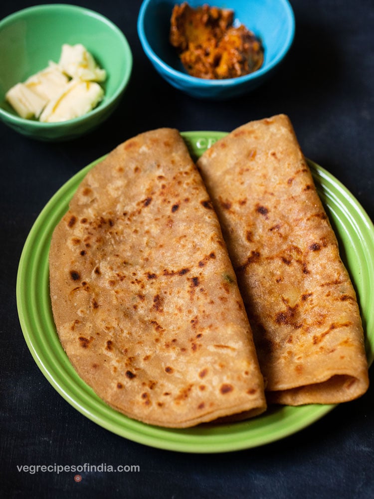 aloo paratha folded and placed on a green ceramic plate on a dark bluish board. sides of butter cubes and mango pickle served in two small ceramic bowls.