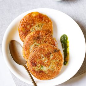 Three aloo tikki kept vertically on a white plate with a spoon and a splash of cilantro dip on the plate placed on a light grey fabric