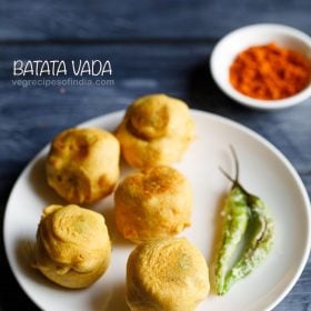 batata vada served on a white plate with a side of with green chilies, a bowl of dry garlic chutney in the background and text layovers.