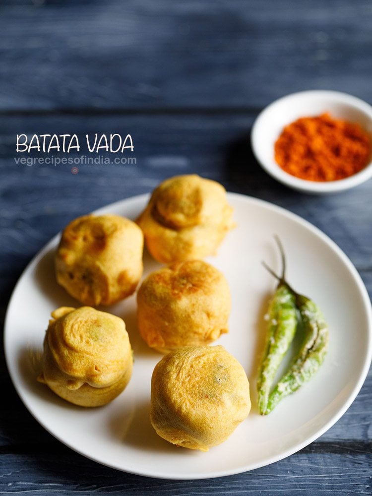 batata vada served on a white plate with a side of with green chilies, a bowl of dry garlic chutney in the background and text layovers.