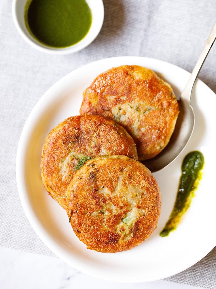 Three aloo tikki kept vertically on a white plate with a spoon and a splash of cilantro dip on the plate placed on a light grey fabric