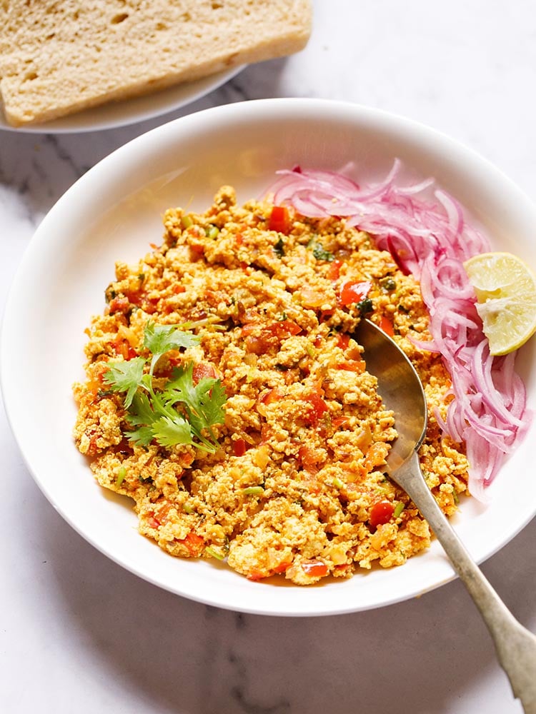 Paneer Bhurji served in a shallow white bowl garnished with one coriander sprig with a spoon inside the bhurji and topped with some sliced onions and a wedge of lemon