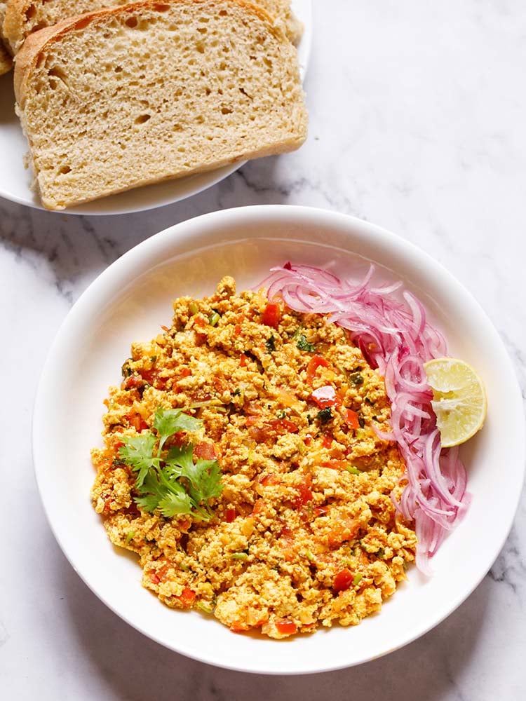 paneer bhurji garnished with a coriander leaf and served on a white plate with onion strips and lemon wedges with a plate of bread slices kept on the top left side. 