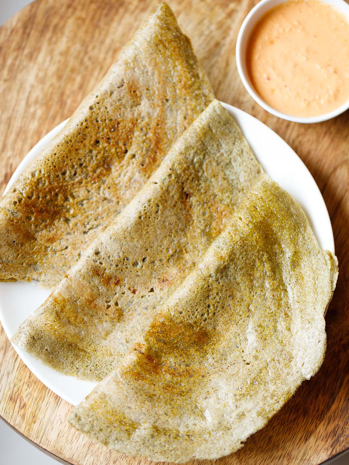 three pesarattu or moong dal dosa folded and kept on a white plate with a orange colored coconut chutney in a side bowl