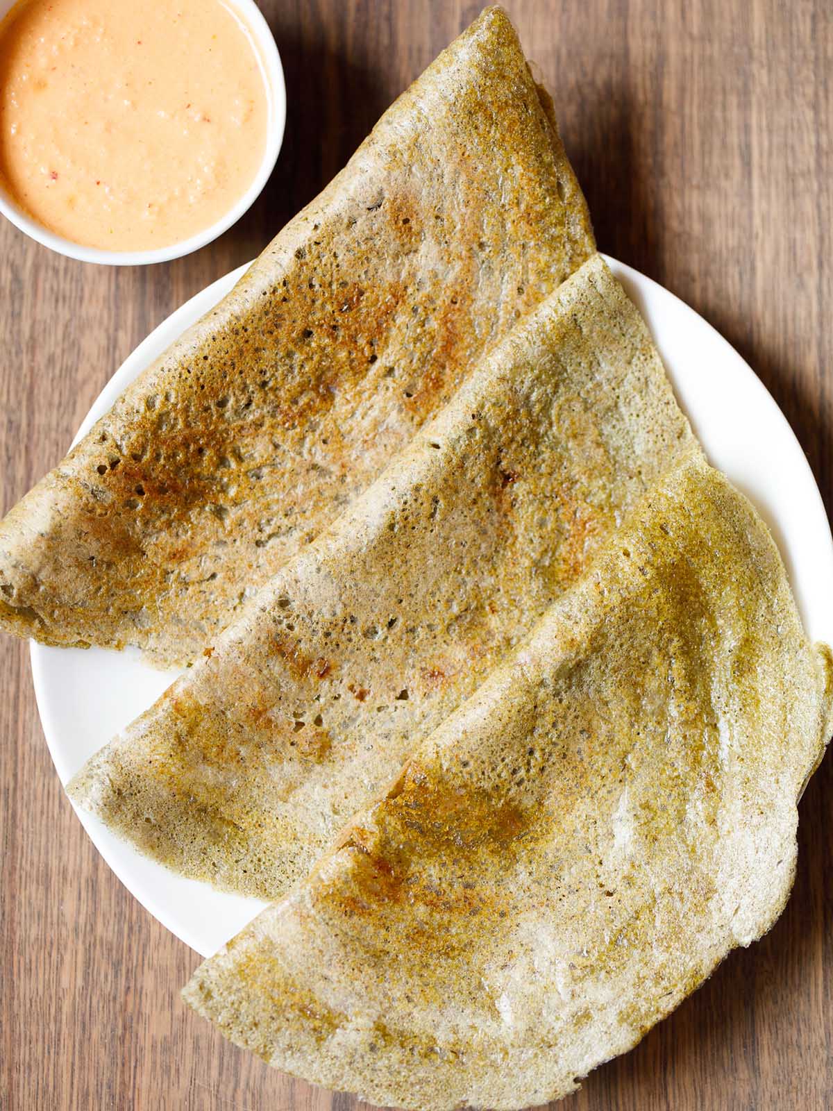 three pesarattu or moong dal dosa folded and kept on a white plate with a orange colored coconut chutney in a side bowl