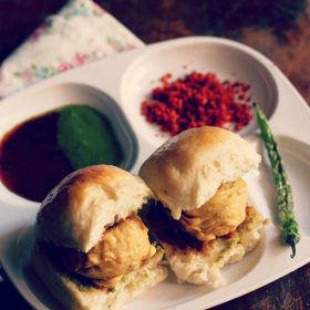 vada pav served with salted green chilies, dry garlic chutney and green chutney on a white plate with sections.