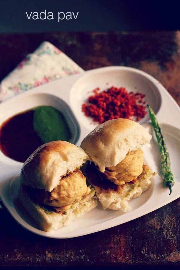 vada pav served with salted green chilies, dry garlic chutney and green chutney on a white plate with sections.