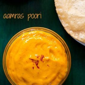 top shot of aamras served in glass bowl with a few saffron strands on top with a side of poori on a green wooden board with a text layover.