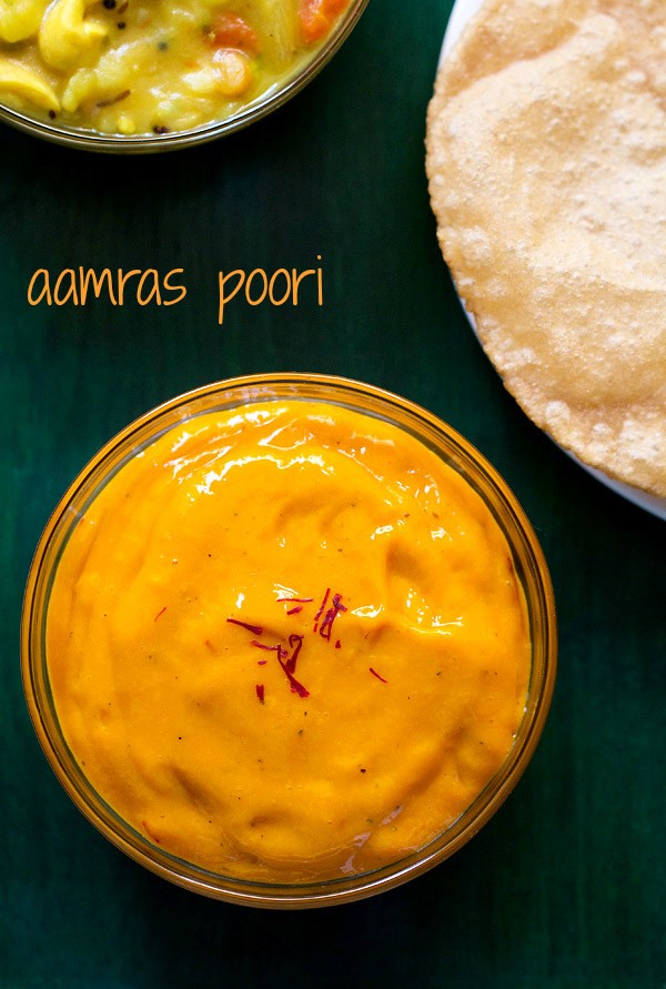 top shot of aamras served in glass bowl with a few saffron strands on top with a side of poori on a green wooden board with a text layover.