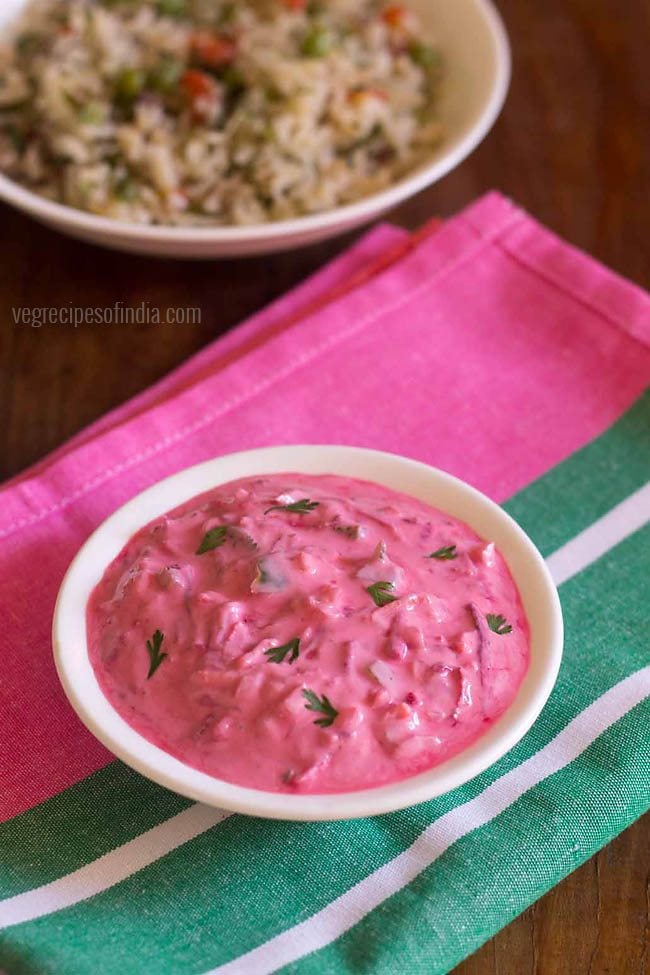 beetroot raita garnished with coriander leaves and served in a white bowl on top of a dark pink and green colored folded cotton napkin.