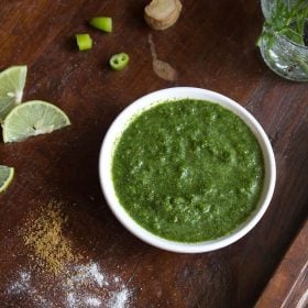 cilantro mint chutney served in white bowl on a wooden board with slices of lemon, chopped green chillies and ginger and sprinkled salt and cumin powder on a wooden board.