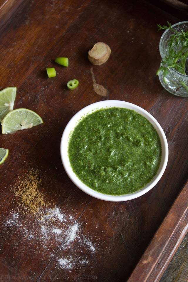 cilantro mint chutney in white bowl on a wooden board with slices of lemon, chopped green chillies and ginger and sprinkled salt and cumin powder on a wooden board