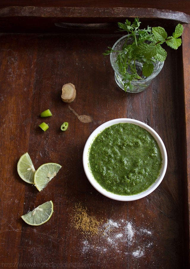 cilantro mint chutney in white bowl on a wooden board with slices of lemon, chopped green chillies and ginger and sprinkled salt and cumin powder on a wooden board