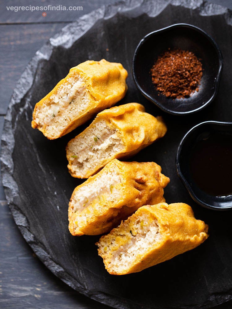 stuffed bread pakoda slices on a black slate board with two black small bowl of dry garlic chutney and tamarind chutney