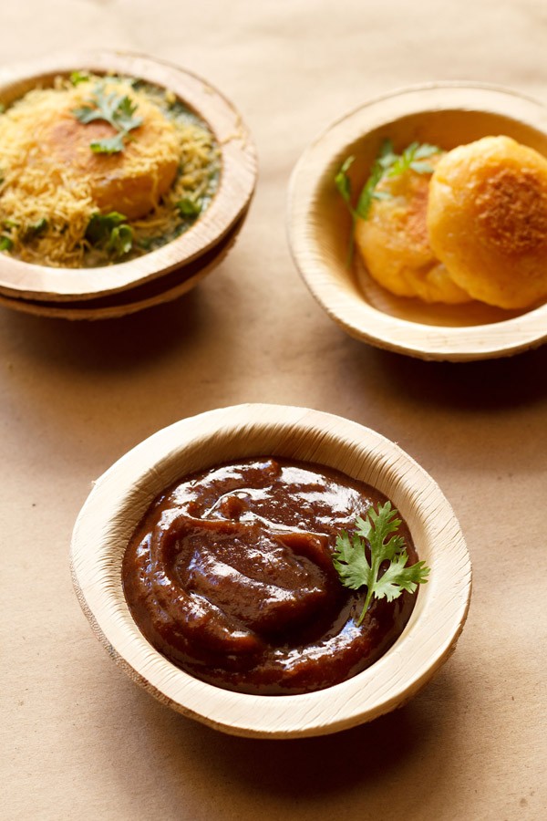 tamarind dates chutney garnished with a coriander sprig and served in an Areca leaf bowl on a light brown paper with potato patties and ragda patties chaat in Areca leaf bowls placed on top 