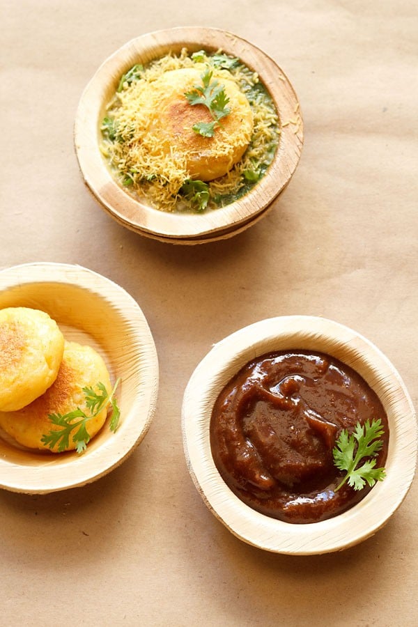 sweet tamarind chutney garnished with a coriander sprig and served in an Areca leaf bowl on a light brown paper with potato patties in areca leaf bowls on left side and ragda patties chaat on top