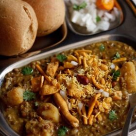 misal served in a plate topped with farsan with a side of sliced lemons and chopped onions and buns.