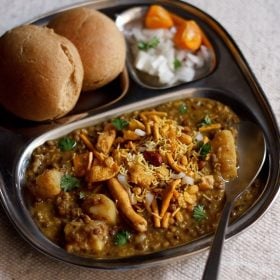 misal served in a plate topped with farsan with a side of sliced lemons and chopped onions and buns.