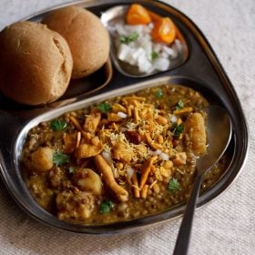 misal served in a plate topped with farsan with a side of sliced lemons and chopped onions and buns