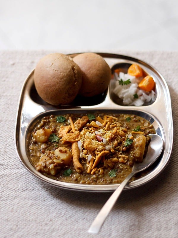 misal served in a plate topped with farsan with a side of sliced lemons and chopped onions and buns