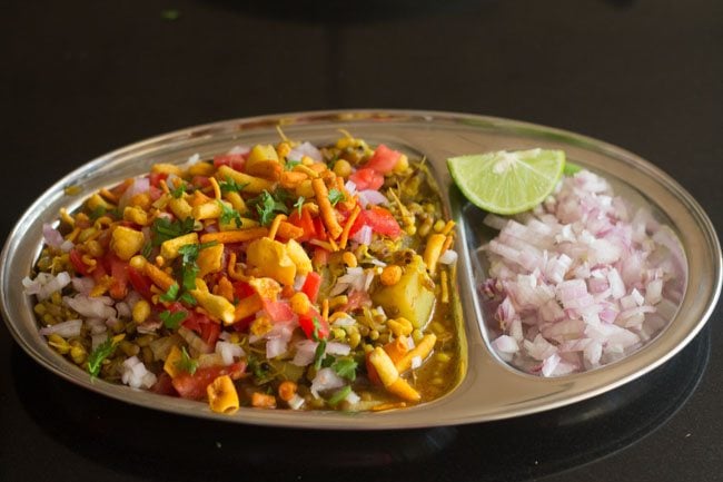 misal served in a plate topped with farsan with a side of sliced lemons and chopped onions