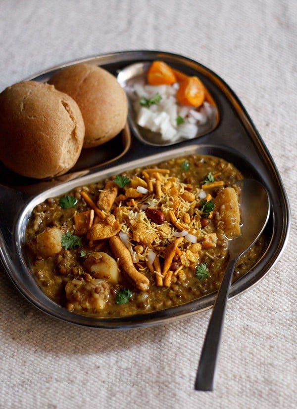 misal served in a plate topped with farsan with a side of sliced lemons and chopped onions and buns