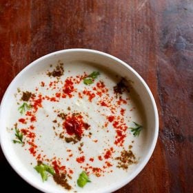 cucumber raita sprinkled with red chilli powder, roasted cumin powder and cilantro in a white bowl on a dark brown wooden board