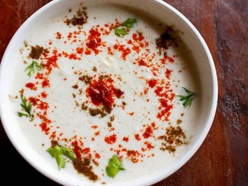 cucumber raita sprinkled with red chilli powder, roasted cumin powder and cilantro in a white bowl on a dark brown wooden board