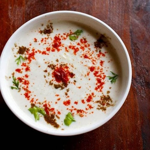 cucumber raita sprinkled with red chilli powder, roasted cumin powder and cilantro in a white bowl on a dark brown wooden board