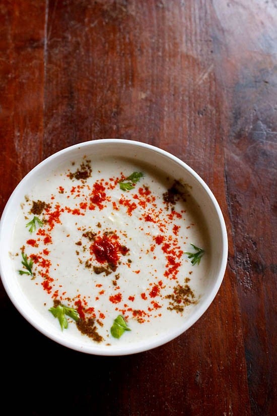cucumber raita sprinkled with red chilli powder, roasted cumin powder and cilantro in a white bowl on a dark brown wooden board