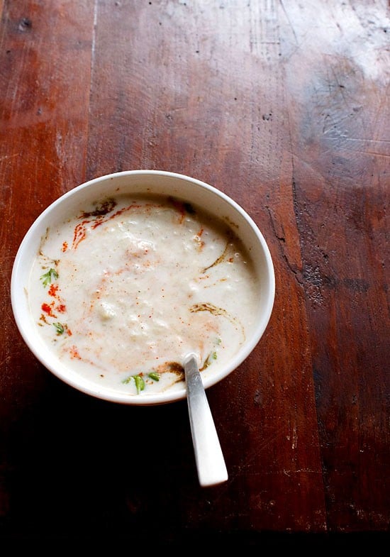 cucumber raita in white bowl with a spoon inside on a dark brown board