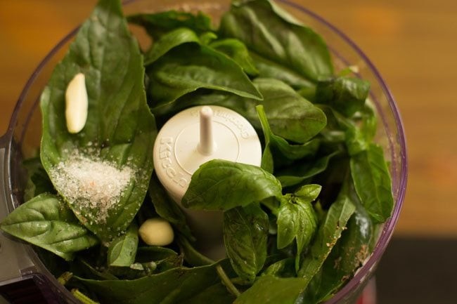 basil, salt, and garlic cloves added to food processor for making vegan pesto sauce recipe.