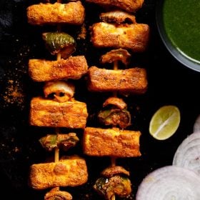 two grilled paneer tikka skewers kept on a black slate board with a bowl of cilantro dip on top right, one lemon wedge below the bowl and a few onion rounds near the lemon wedge