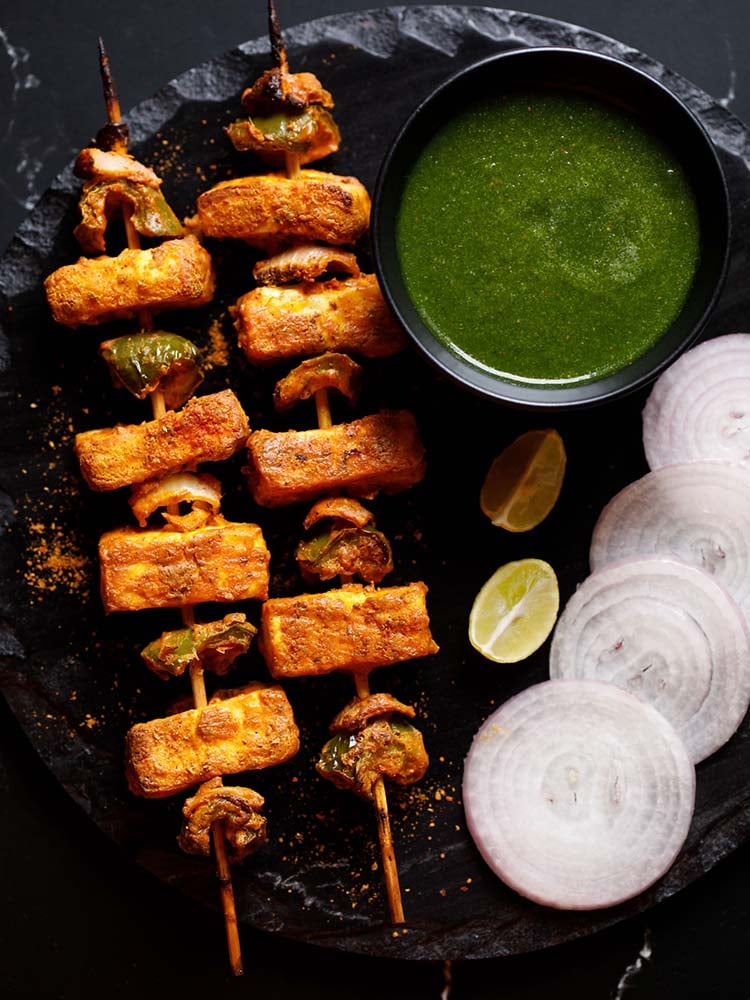 two grilled paneer tikka skewers kept on a black slate board with a bowl of cilantro dip on top right, two lemon wedges below the bowl and some slices onion rounds near the lemon wedges
