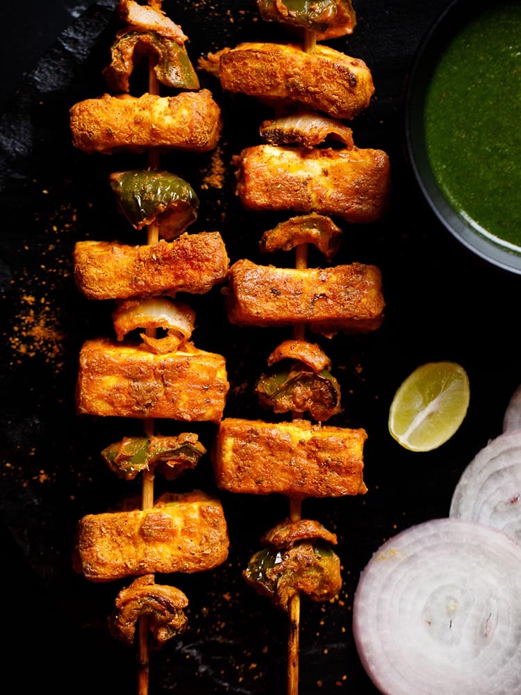 two grilled paneer tikka skewers kept on a black slate board with a bowl of cilantro dip on top right, one lemon wedge below the bowl and a few onion rounds near the lemon wedge
