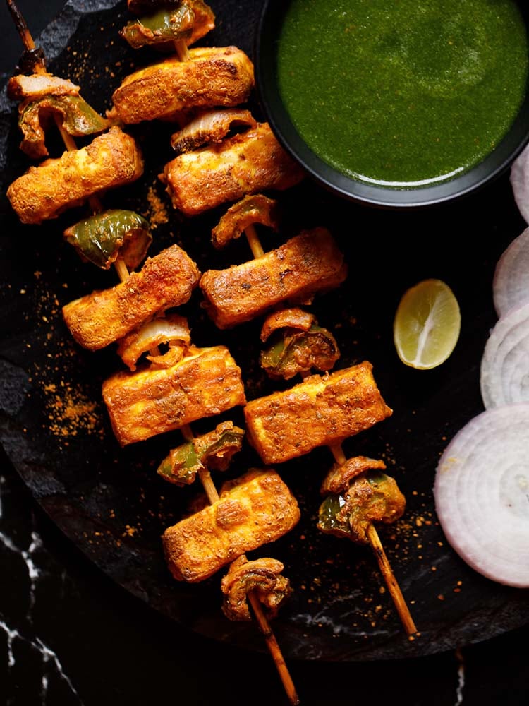 wo grilled paneer tikka skewers kept on a black slate board with a bowl of cilantro dip on top right, one lemon wedge below the bowl and a few onion rounds near the lemon wedge