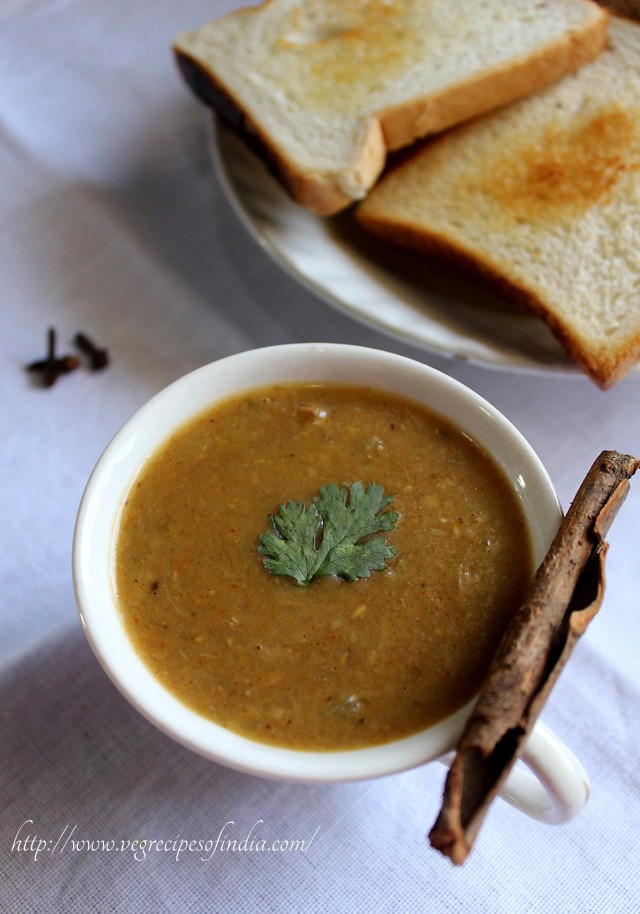 veg dhansak garnished with coriander leaf and served in a large white mug with a cinnamon kept on the side and toasted bread on a white plate on the side. 