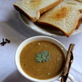 dhansak garnished with coriander leaves and served in a white bowl with toasted bread kept on a plate in the side.