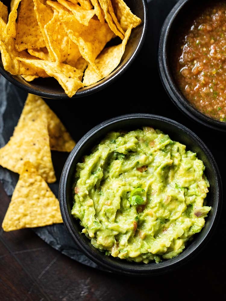 guacamole served in a black wooden bowl with a side black bowl of nachos and tomato salsa