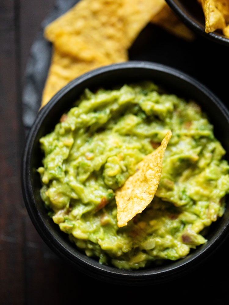 tortilla chip dipped in guacamole dip in a wooden black bowl on a round black granite board.