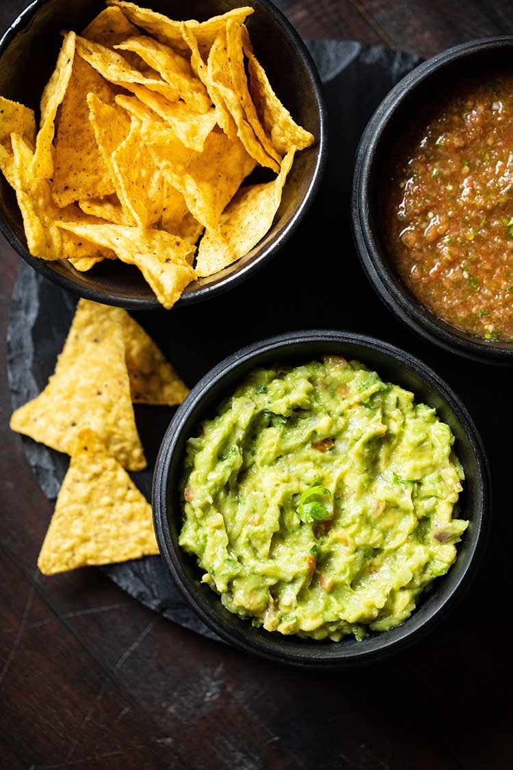 avocado served in a black wooden bowl with a side black bowl of tortilla chips and tomato salsa.