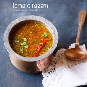 Tomato rasam in a traditional South Indian bowl with a spoon on a white kitchen napkin placed left on a blue board