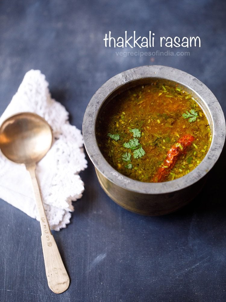 thakkali rasam in a traditional South Indian container with a spoon placed on a white doily napkin next to the container on a dark blue board