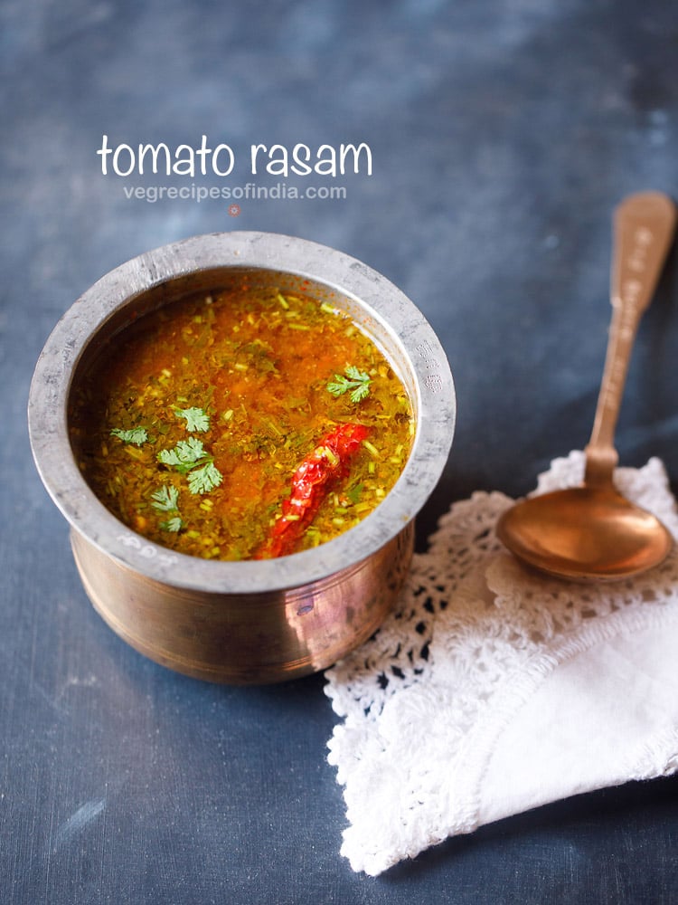 tomato rasam in a traditional South Indian container with a spoon placed on top of a white kitchen napkin on the left side on a blue board