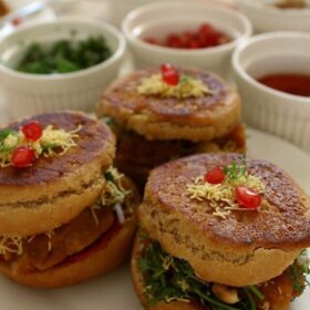 side shot of dabeli served on a white plate with garnished kept in white bowls on the upper side.