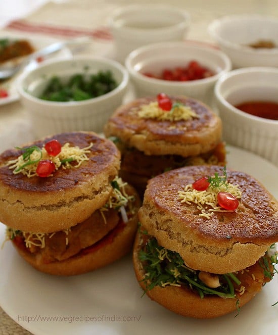 side shot of dabeli served on a white plate with garnished kept in white bowls on the upper side.